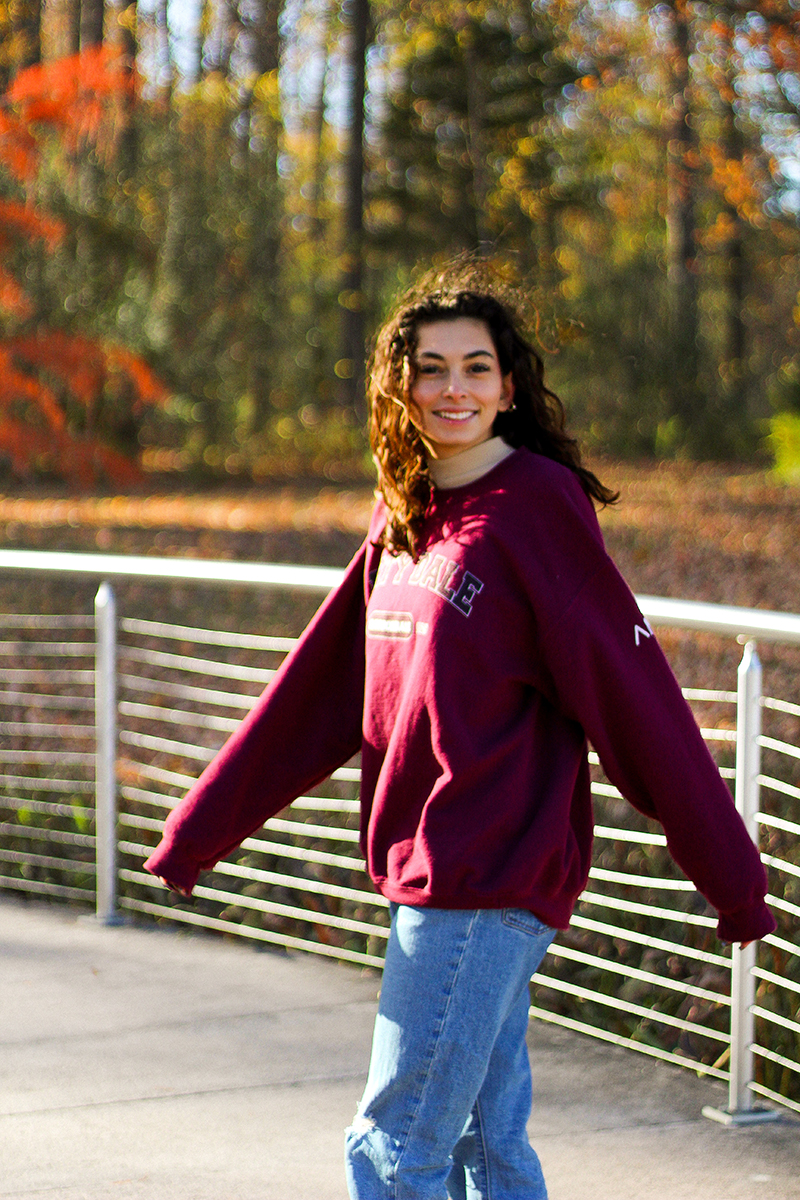 Trinity is smiling while wearing a maroon SIU sweatshirt and blue jeans walks along a metal-railed bridge, with colorful autumn trees and sunlight in the background.