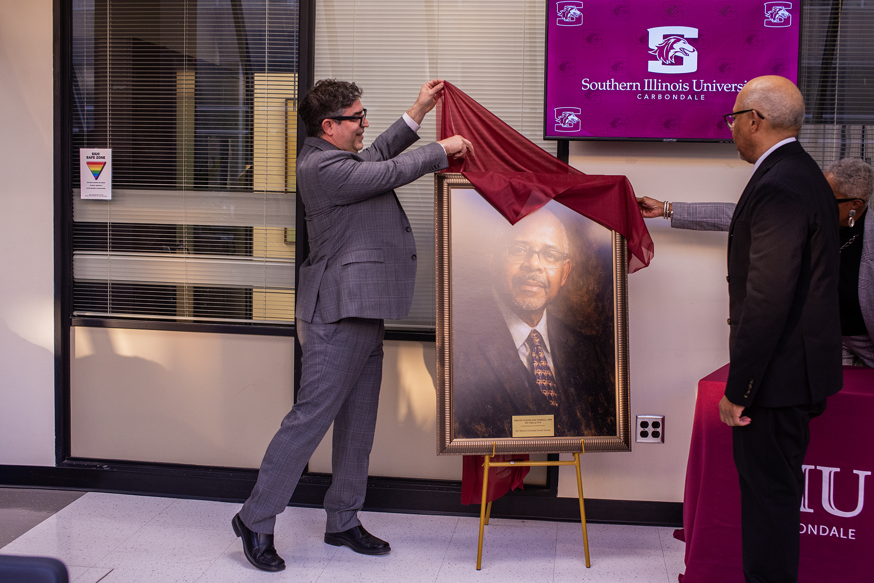 Dr. Benjamin Bricker, acting dean of the College of Liberal Arts at Southern Illinois University Carbondale, unveils a framed portrait of Dr. Melvin C. Terrell during a dedication ceremony. Bricker lifts a maroon cloth to reveal the portrait, which is displayed on an easel, as attendees look on. A screen behind them displays the Southern Illinois University Carbondale logo.