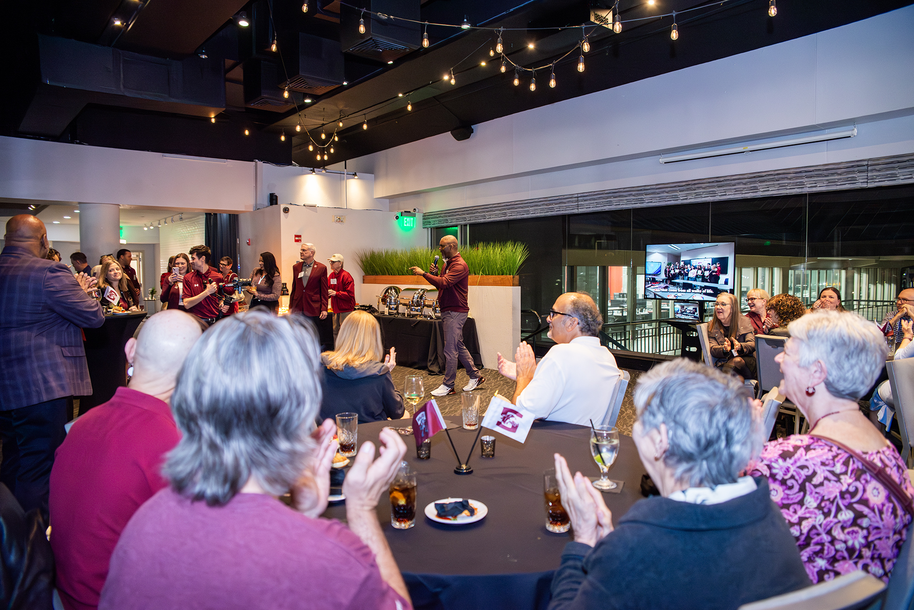 SIU Chancellor Austin A. Lane speaks to alumni and friends during the Chancellor’s Reception in Tampa as attendees seated at round tables applaud, with SIU flags on the tables and a city view visible through the windows behind him.