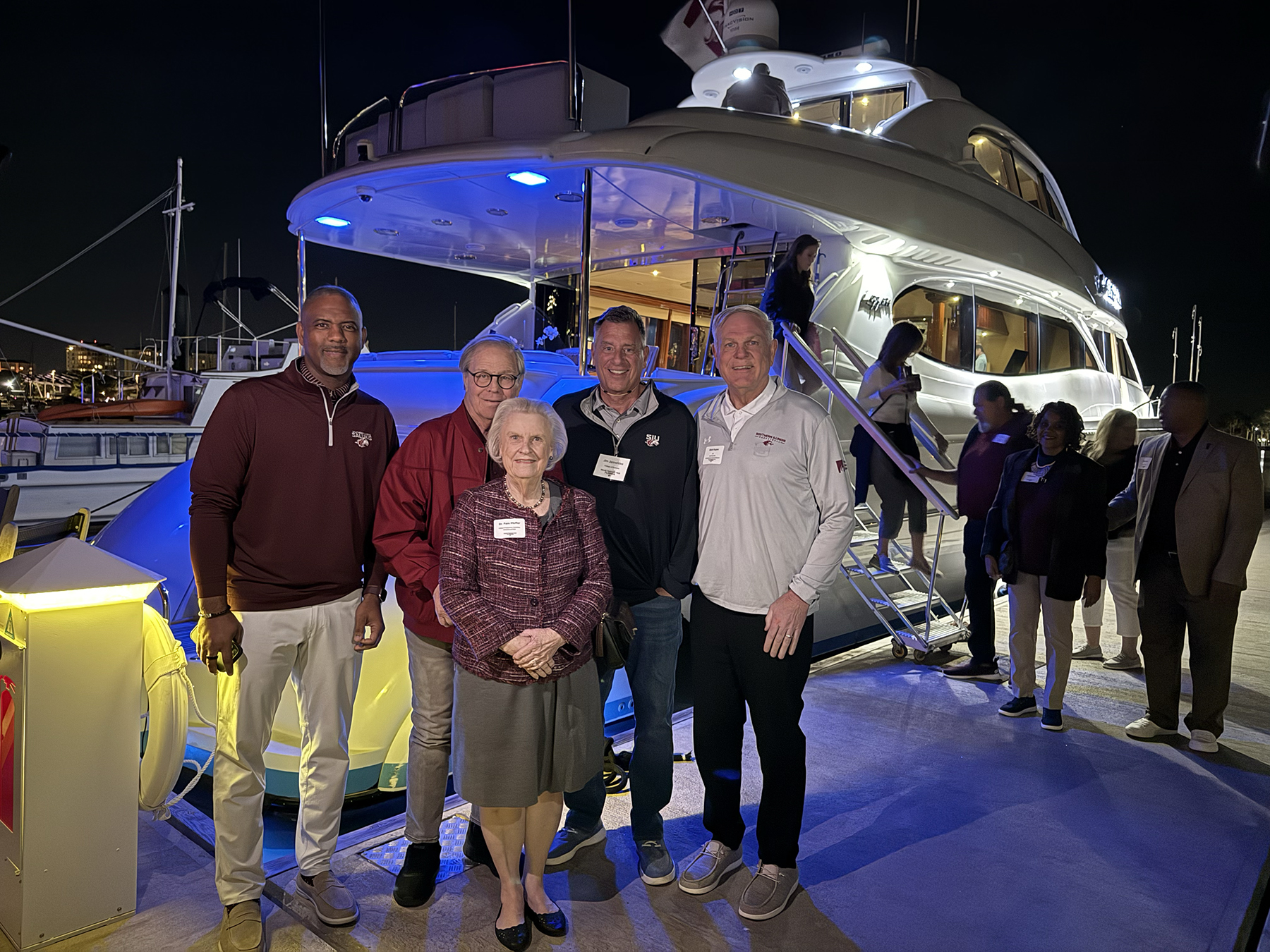 SIU Chancellor Austin Lane, Dr. Phil Pfeffer, Dr. Pam Pfeffer, James Jannetides and SIU Foundation CEO Matt Kupec stand together on a marina dock at night in front of a lit yacht during the Saluki Takeover Tour Florida kickoff event in St. Petersburg.