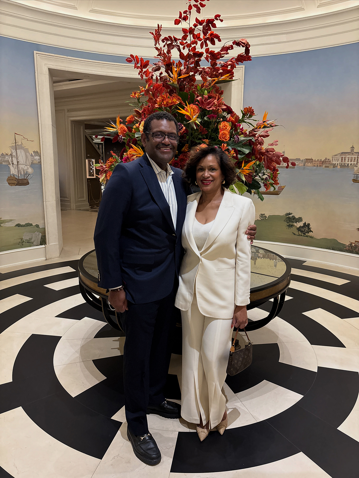 Raymond and Lisa Hankins stand together smiling indoors, dressed in formal attire, in front of a tall autumn floral arrangement with red and orange leaves.