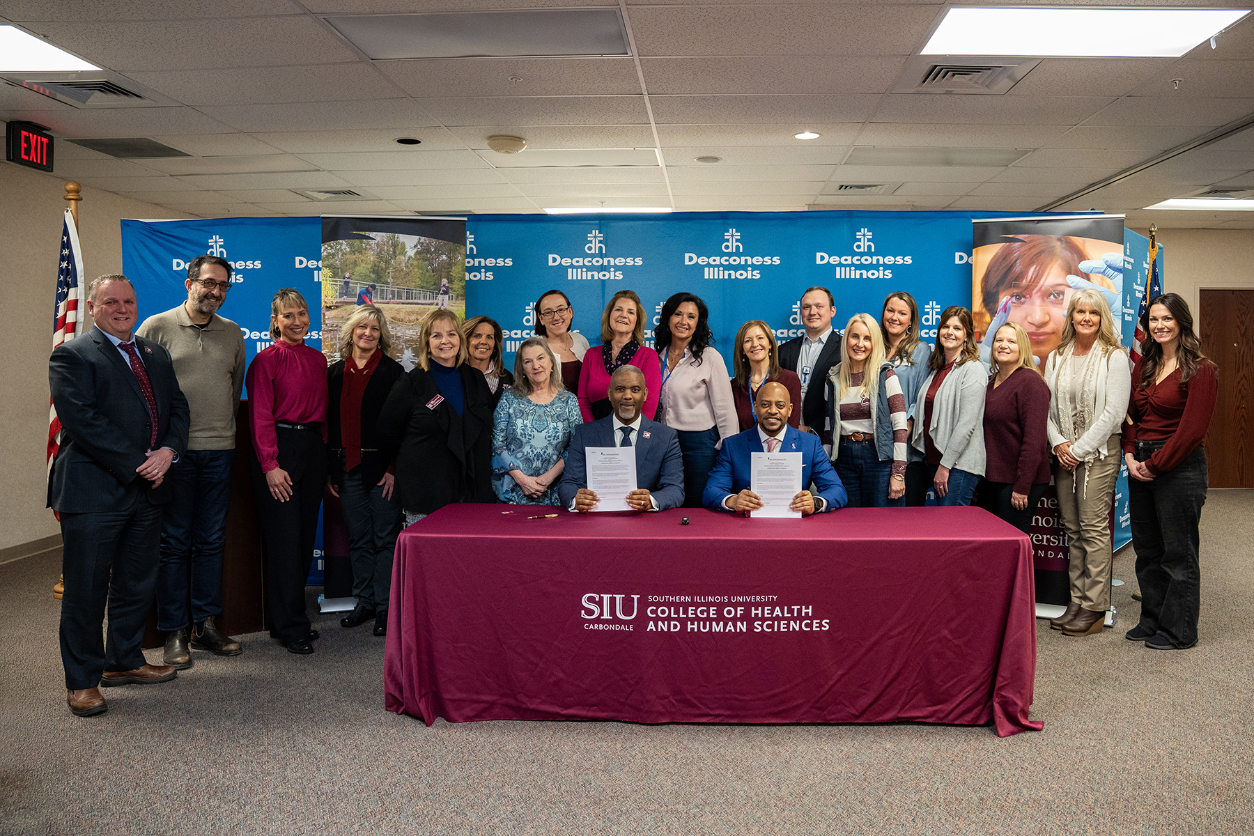 SIU Chancellor Austin A. Lane, seated left, and William Davis, Deaconess Illinois Region President, sign partnership agreements at a table draped with an SIU College of Health and Human Sciences tablecloth Tuesday, Feb. 24, 2026, at Deaconess Illinois Medical Center in Marion. Leaders and team members from Deaconess Illinois and the college stand behind them in front of a Deaconess Illinois backdrop.