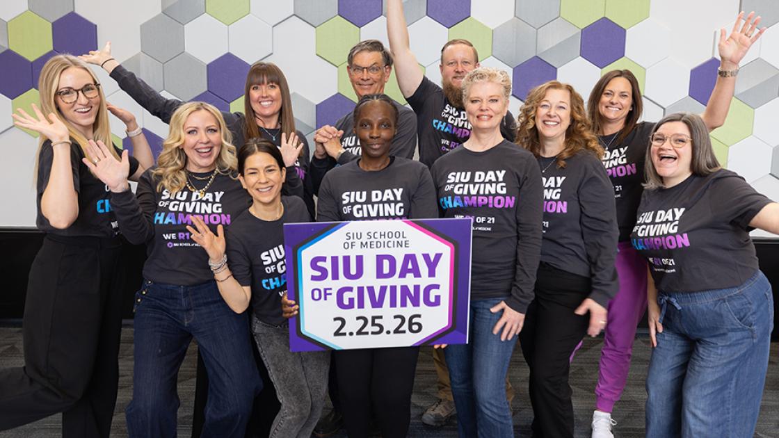 Group of people wearing matching “SIU Day of Giving Champion” shirts pose energetically in front of a geometric wall, holding a sign that reads “SIU School of Medicine – SIU Day of Giving – 2.25.26.”