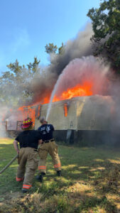 Firefighters spray water onto a mobile home engulfed in flames during an active structure fire response.
