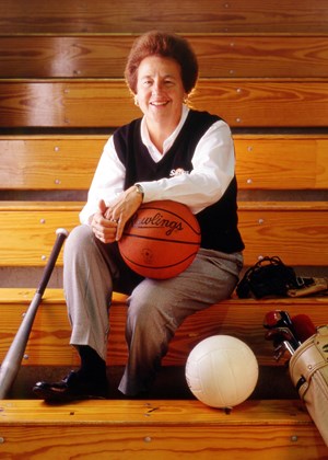 Dr. Charlotte West sits on wooden gym bleachers holding a basketball, smiling at the camera. She wears a white shirt and dark vest with an SIU logo. A volleyball, baseball bat and glove rest beside her, symbolizing the multiple sports she coached and supported during her career in women’s athletics.
