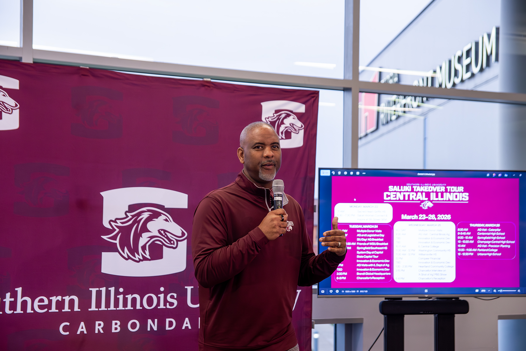 Chancellor Austin A. Lane speaks to attendees during the Saluki Takeover Tour Chancellor’s Reception at the Peoria Riverfront Museum, standing in front of an SIU backdrop and event display.
