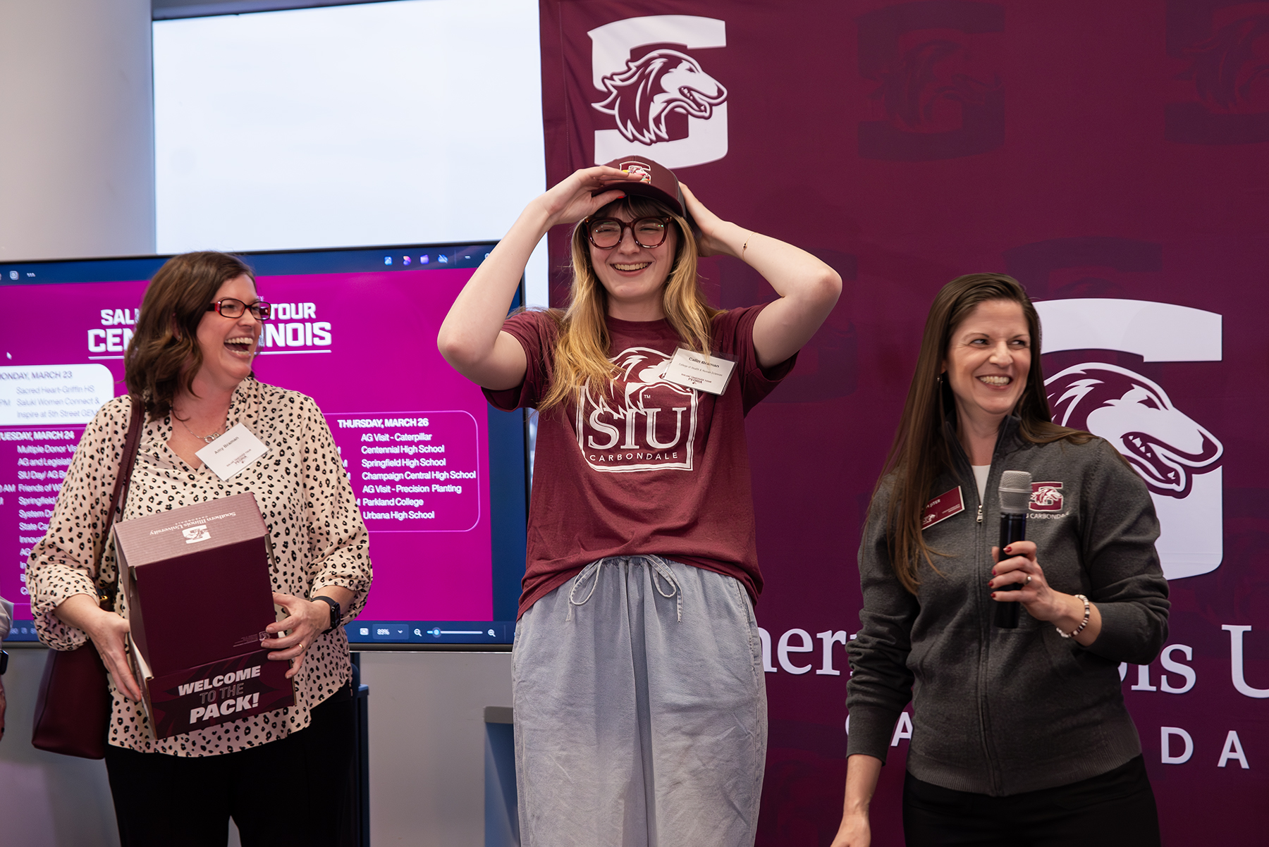 Incoming SIU student Calin Braman smiles while trying on an SIU hat during a recognition moment at the Saluki Takeover Tour Chancellor’s Reception in Peoria, joined by Sarah Jiter, director of undergraduate admissions, and her mom.