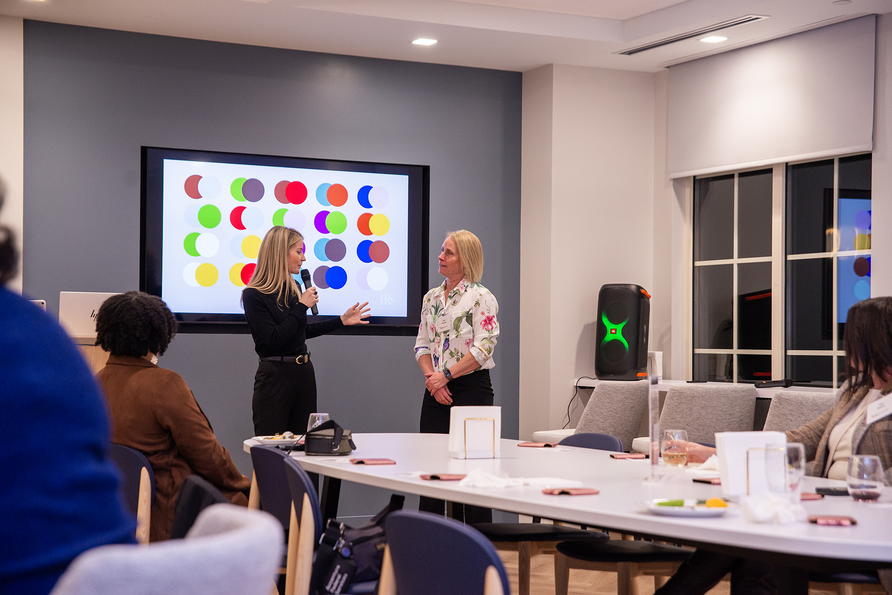 Laura Sawyier, founder and CEO of LKS Styling and Consulting, speaks into a microphone during the Saluki Women Connect and Inspire event at Moneta in Clayton, Missouri, while another attendee stands beside her as guests listen from tables in the room.