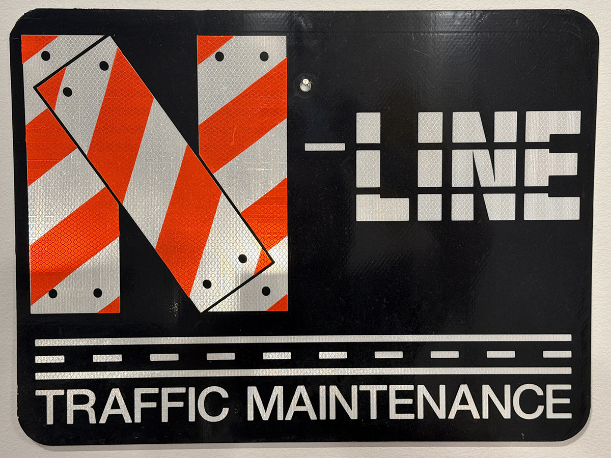 N-Line Traffic Maintenance logo - Black sign with reflective red-and-white diagonal stripes forming a large letter “N,” the words “N-Line” and “Traffic Maintenance,” and a stylized roadway graphic along the bottom.