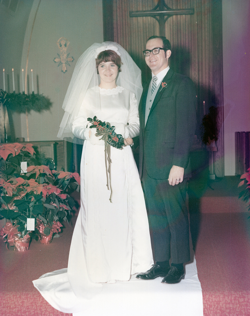 Pat and Bob England are standing together at the front of a church on their wedding day, wearing formal wedding attire, with floral arrangements, candles, and a cross visible in the background.