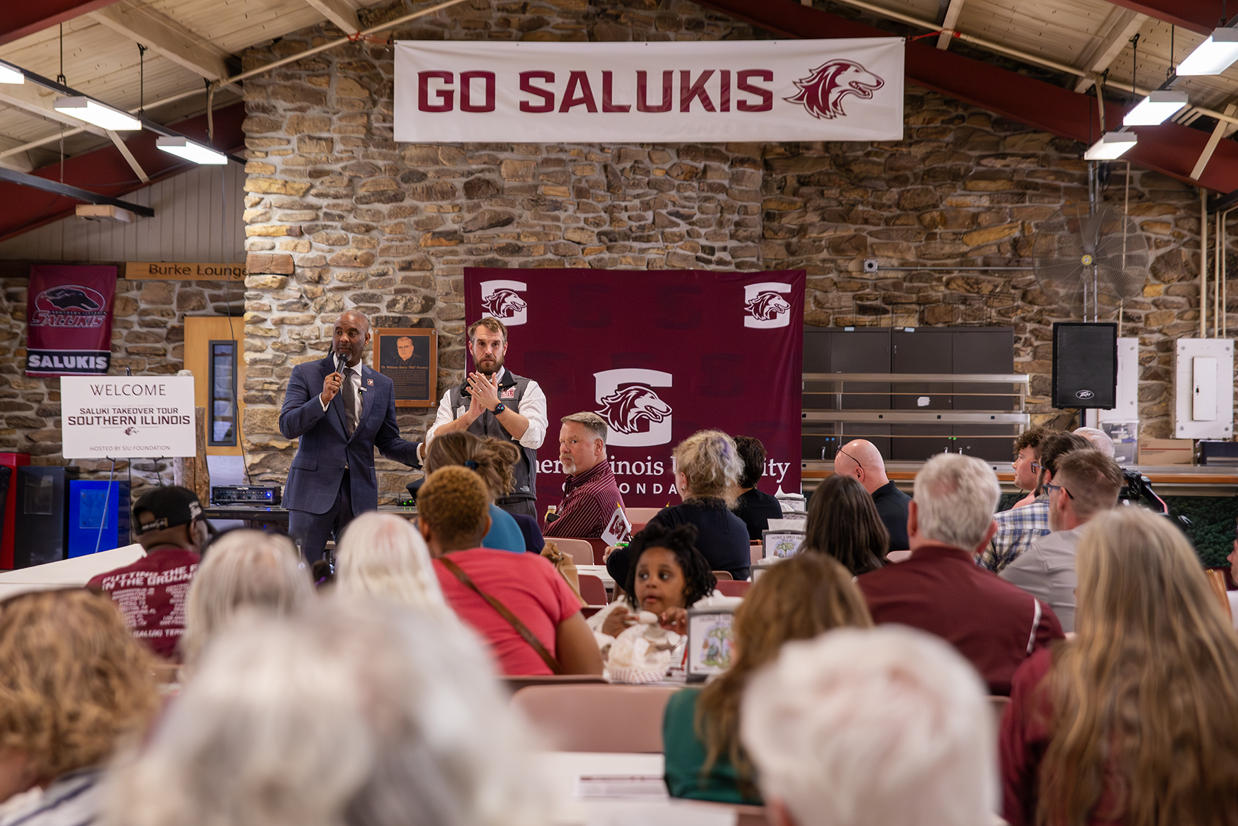 Chancellor Austin A. Lane speaks to a crowd of alumni, students and community members while Brian Croft stands beside him during the kickoff of the Saluki Takeover Tour Southern Illinois at Touch of Nature Outdoor Education Center, with a “Go Salukis” banner displayed above the stage.