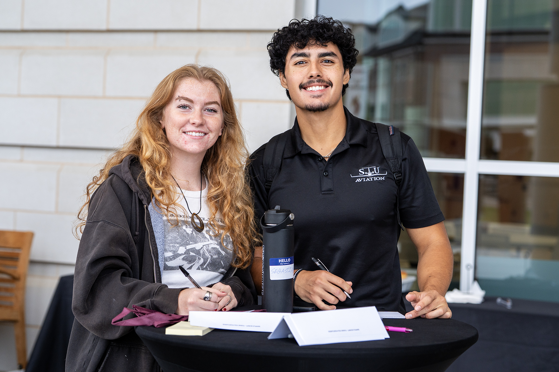 Photo of female and male student smiling at the camera while writing a thank-you note to donors on a postcard.
